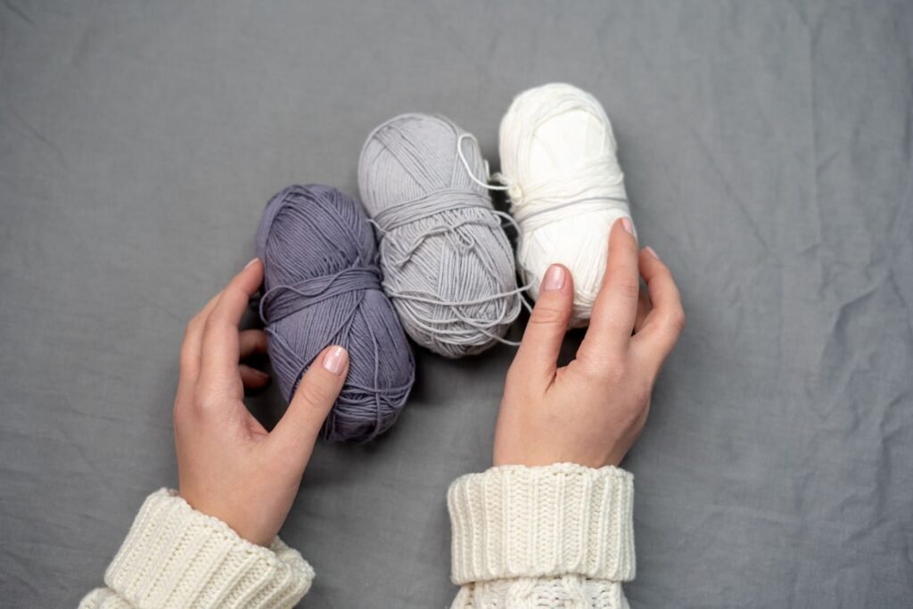 Cozy knitting setup with hands holding purple, gray, and white yarn balls on a soft surface.