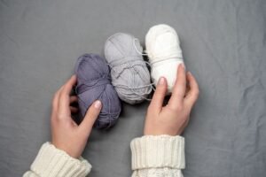 Cozy knitting setup with hands holding purple, gray, and white yarn balls on a soft surface.