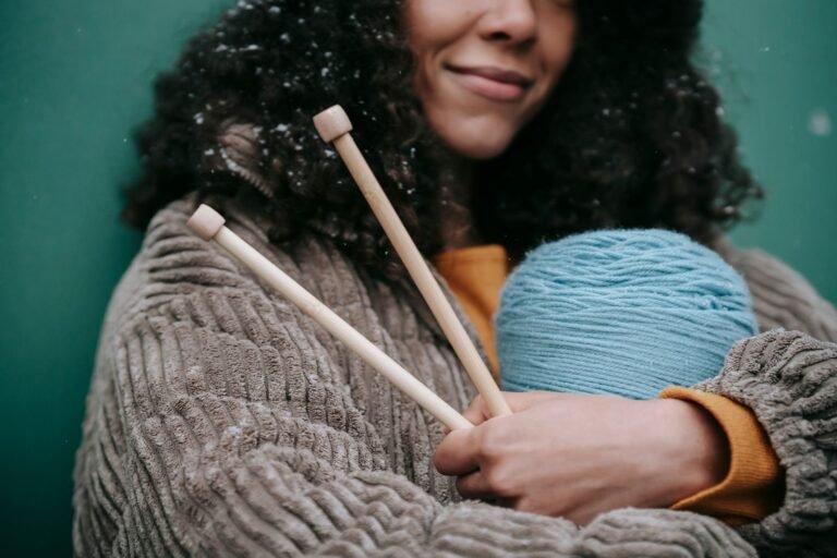 Crop smiling African American woman with snowflakes on dark curly hair wearing beige corduroy jacket cuddling skein of blue yarn and knitting needles